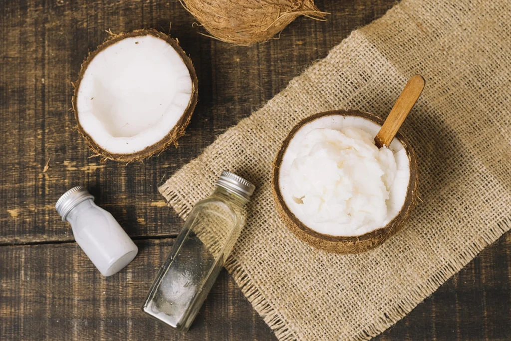 Halved coconuts with one filled with solid coconut oil and a wooden spoon, next to small bottles of clear and white liquids, arranged on a wooden surface with burlap fabric.