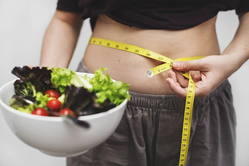 A person measuring their waist with a tape measure while holding a bowl of fresh salad with cherry tomatoes and leafy greens.