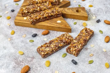 Granola-style protein bars with oats, seeds, nuts, and a chocolate drizzle, displayed on a wooden board and surrounded by loose ingredients.