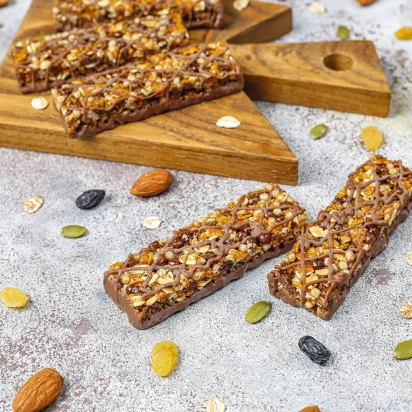 Granola-style protein bars with oats, seeds, nuts, and a chocolate drizzle, displayed on a wooden board and surrounded by loose ingredients.