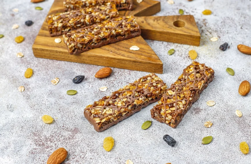 Granola-style protein bars with oats, seeds, nuts, and a chocolate drizzle, displayed on a wooden board and surrounded by loose ingredients.