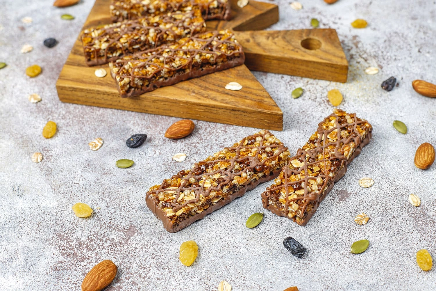 Granola-style protein bars with oats, seeds, nuts, and a chocolate drizzle, displayed on a wooden board and surrounded by loose ingredients.