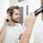 Man giving himself a haircut with electric clippers while smiling in front of a bathroom mirror.