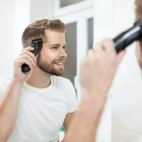 Man giving himself a haircut with electric clippers while smiling in front of a bathroom mirror.