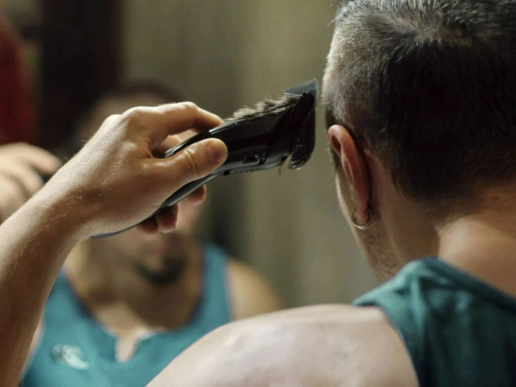 Man using hair clippers to cut his own hair to sides of his head while looking in the mirror.