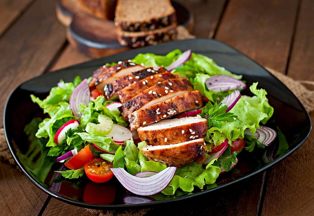 A bowl of grilled chicken slices, boiled egg halves, broccoli, cauliflower, cherry tomatoes, avocado, and grilled pineapple, served with rustic bread and fresh tomatoes.