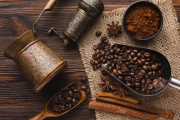 Traditional coffee setup with roasted coffee beans, ground coffee, spices, and antique brass utensils on a rustic wooden table
