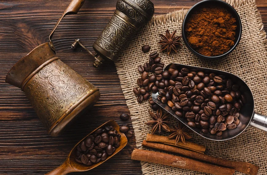 Traditional coffee setup with roasted coffee beans, ground coffee, spices, and antique brass utensils on a rustic wooden table