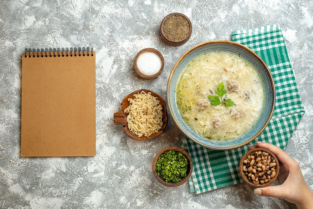 Top view of a rustic kitchen setup with a bowl of creamy soup containing meatballs and herbs, surrounded by ingredients like noodles, black-eyed peas, chopped parsley, salt, and pepper, alongside a blank notepad on a textured gray surface.