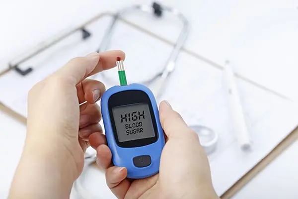 A person holding a blue blood glucose meter showing a 'HIGH BLOOD SUGAR' warning, with a test strip inserted and a drop of blood on the tip. Medical tools, including a stethoscope and clipboard, are visible in the background.