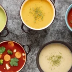 An overhead view of five colorful bowls of soup, including creamy green broccoli soup, yellow squash soup, rich tomato soup with herbs, hearty vegetable soup with carrots and broccoli, and creamy potato soup garnished with herbs, displayed on a textured gray surface