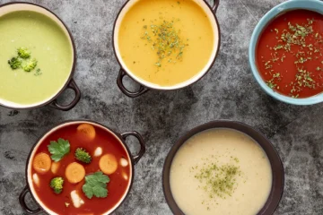 An overhead view of five colorful bowls of soup, including creamy green broccoli soup, yellow squash soup, rich tomato soup with herbs, hearty vegetable soup with carrots and broccoli, and creamy potato soup garnished with herbs, displayed on a textured gray surface