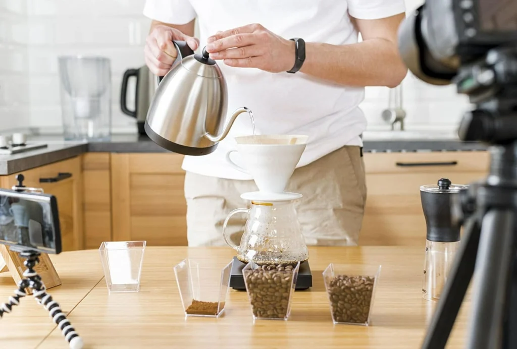 Person making pour-over coffee with a stainless steel kettle in a modern kitchen, surrounded by coffee beans and recording equipment.