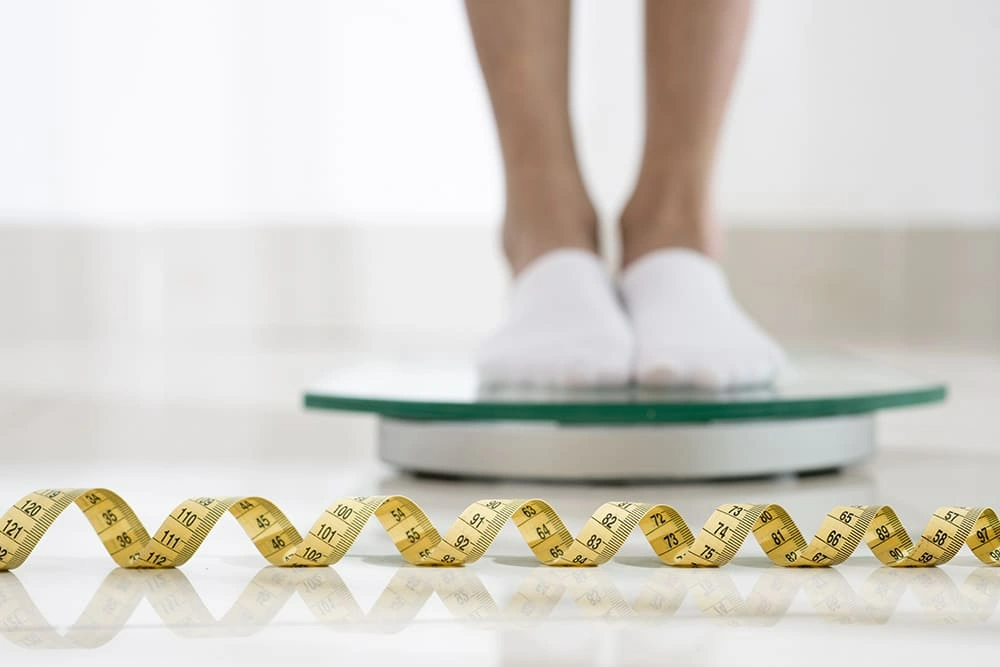 Person standing on a digital bathroom scale with a yellow measuring tape on the floor, symbolizing weight loss and body measurement tracking.