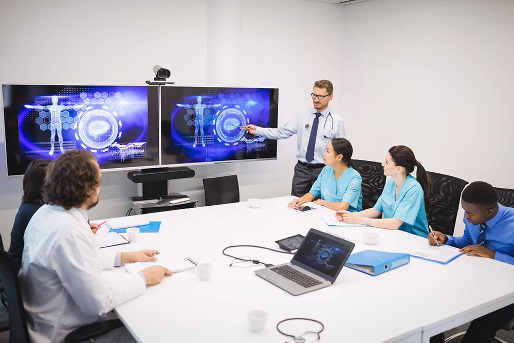 Doctor presenting medical data on dual screens to a group of healthcare professionals during a team meeting